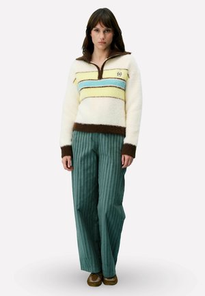 Vrouw die een witte, pluizige trui draagt met blauwe en gele strepen, groene broek met wijde pijpen en bruine mocassin-achtige schoenen.