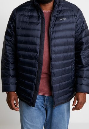 Homme portant une veste matelassée bleu marine Calvin Klein sur une chemise bordeaux et un jean bleu clair, debout les mains détendues le long du corps.