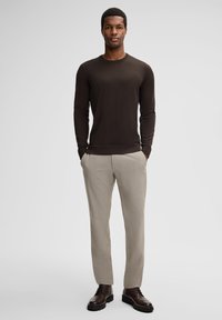 Ein Mann steht mit den Händen in den Taschen, trägt einen dunkelbraunen Pullover, beigefarbene Hosen und dunkelbraune Lederstiefel vor einem einfarbigen Hintergrund.