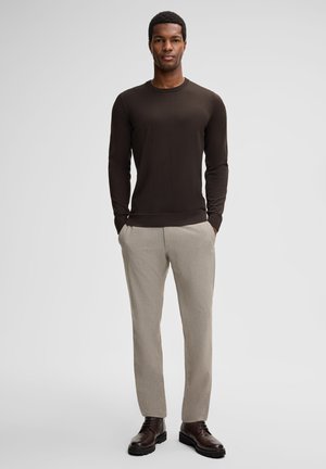 Man stands with hands in pockets wearing dark brown sweater, beige trousers, and dark brown leather boots against a plain background.