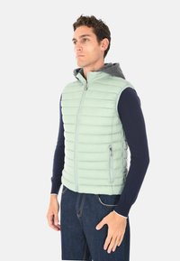 Uomo che indossa un gilet smanicato trapuntato verde chiaro con zip e cappuccio grigio sopra una maglietta a maniche lunghe blu navy e jeans blu, rivolto a sinistra.