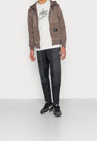 Vaaleanruskea vetoketjullinen huppari etutaskuilla, yhdistettynä mustiin kevyisiin housuihin, joissa on vetoketjullinen tasku ja graafisia yksityiskohtia. Mustat tennarit täydentävät asukokonaisuuden.