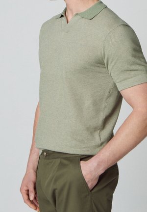 Jasnozielona, teksturowana koszulka polo z krótkim rękawem, dekoltem w kształcie litery V i kontrastowym kołnierzykiem, w połączeniu z ciemnozielonymi spodniami. Brak widocznych wzorów lub akcentów.