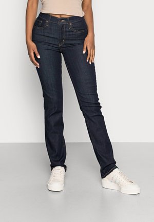 Dark blue jeans with a straight leg design, featuring five pockets and copper stitching, paired with white sneakers on a gray floor.