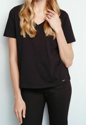 Mujer rubia con una camisa negra lisa de manga corta y cuello en V y pantalones negros, de pie frente a un fondo claro.