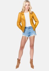 Žlutá kožená motocyklová bunda se zipovými uzávěry, nošená přes bílou košili, doplněná o nařasené džíny a hnědé kotníkové boty.