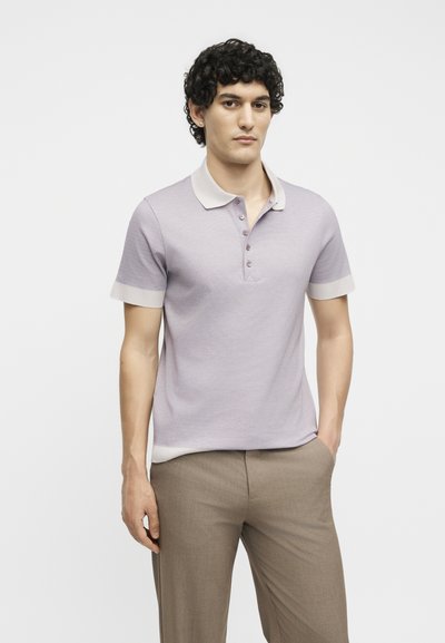 Νέος άνδρας με σγουρά σκούρα μαλλιά, που φοράει ανοιχτό μοβ πόλο μπλουζάκι με μπεζ γιακά και μανσέτες στα μανίκια και μπεζ παντελόνι, στέκεται με το ένα χέρι στην τσέπη.