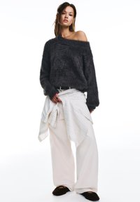Młoda kobieta stoi ubrana w puszysty, ciemny sweter z odsłoniętymi ramionami, szerokie białe spodnie, jasną koszulę w paski zawiązaną w talii oraz ciemne kapcie.