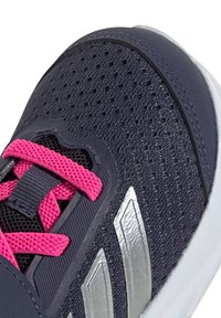 Zapatilla deportiva de color navy con parte superior de malla perforada, cordones rosas y acentos plateados. Presenta superposiciones texturizadas y una suela de goma blanca.