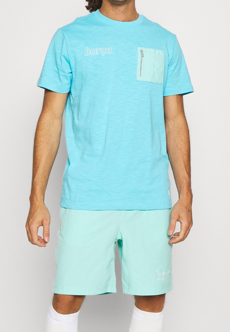 T-shirt bleu clair avec le texte "barça", doté d'une poche avant avec une fermeture éclair. Associé à un short vert menthe affichant une broderie "barça".