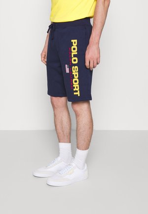 Mann trägt marineblaue Shorts mit Kordelzug, gelbem "POLO SPORT" und rotem "Ralph Lauren" Schriftzug sowie einer amerikanischen Flagge, kombiniert mit weißen Sneakers und Socken.