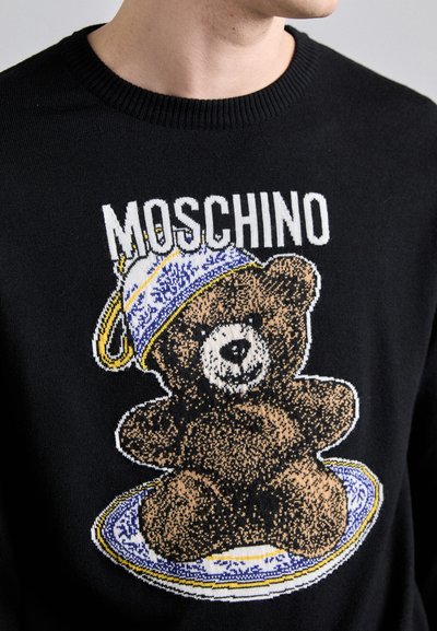 Pulover negru cu un grafic tricotat al unui urs purtând o pălărie albastră și aurie, așezat pe o farfurie, cu textul "MOSCHINO" deasupra designului.