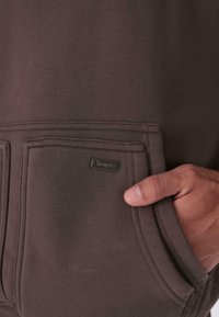 Kéz a barna kapucnis pulcsi elülső zsebében, látható varrással és egy kis fémből készült márkajellel, amely "Dropsia" feliratú.