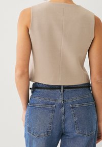 Beżowy top bez rękawów z prostym dołem, wykonany z gładkiej tkaniny, połączony z wysokim stanem niebieskich dżinsów z tylnymi kieszeniami.