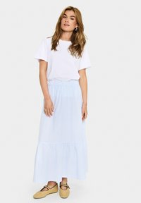 Tricou alb cu mânecă scurtă, asortat cu o fustă maxi cu dungi albastre și albe, având un tiv volanat, purtată cu balerini împletiți de culoare bej.