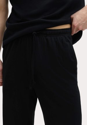 Persona che solleva leggermente una maglietta nera, indossa pantaloni neri con elastico in vita e coulisse, con pelle visibile in vita su sfondo bianco.