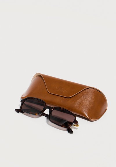 Ορθογώνια γυαλιά ηλίου με μοτίβο χελώνας και σκουρόχρωμους φακούς τοποθετημένα μπροστά από καφέ δερμάτινη θήκη γυαλιών ηλίου σε ανοιχτόχρωμη επιφάνεια.
