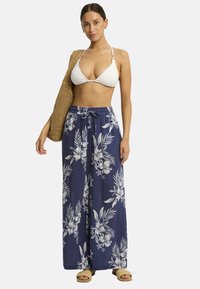 Model in een witte bikini-top en marineblauwe wijdvallende broek met bloemenpatroon, met een geweven tas, staand voor een eentonige achtergrond.