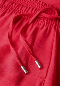 Pantaloni corti rossi in tessuto con vita arricciata, dotati di una coulisse con estremità in metallo argentato. La texture è liscia e leggera.