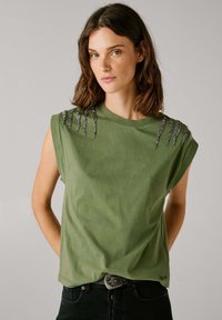 Camiseta sin mangas de color verde oliva con un escote redondo, que presenta acentos de tachuelas plateadas en los hombros. Material de algodón suave.