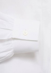 Valkoinen kauluspaita hihansuussa, jossa on pleatattu rakenne ja pyöreä valkoinen nappi, jossa on kukkakuvio. Tasainen kangas ja siisti ommel.