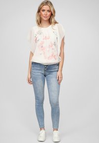 Blusa leggera e trasparente con stampa floreale in rosa chiaro e grigio, abbinata a jeans aderenti azzurri e sneakers bianche.
