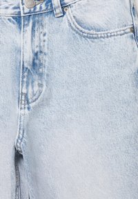 Gros plan d'un jean en denim bleu clair montrant la poche avant, le bouton en métal, la boucle de ceinture et les détails de couture visibles.