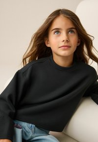 Schwarzer Pullover mit hohem Kragen und überschnittenen Schultern, aus einem glatten Stoff gefertigt. Kombiniert mit hellblauen Jeans mit floralem Akzent.