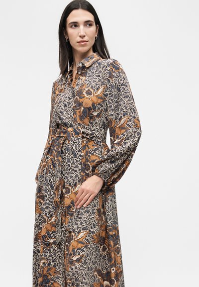 Γυναίκα φοράει μακρυμάνικο φόρεμα με γιακά, με καφέ, πορτοκαλί και μαύρο λουλουδάτο και αφηρημένο σχέδιο, ποζάροντας με το ένα χέρι στη μέση της.