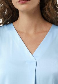 Helles blaues Blusenshirt mit V-Ausschnitt. Glatte Textur, dezent glänzend und mit einer genähten Detail am Mittelpunkt. Nahaufnahme des Ausschnitts und der Schultern.