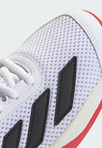 Chaussure de sport blanche avec une tige en maille, des accents noirs et une semelle rouge. Présente des lacets plats et une surface texturée pour une meilleure adhérence.