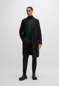 Zwarte double-breasted overjas, gecombineerd met een groene coltrui en zwarte tapered broek. Het model draagt zwarte enkellaarsjes. Soepele textuur, perfecte pasvorm.