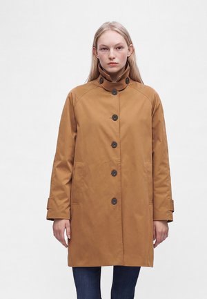 Donna con capelli biondi lisci che indossa un cappotto marrone chiaro abbottonato con collo alto e pantaloni scuri, in piedi davanti a uno sfondo bianco semplice.
