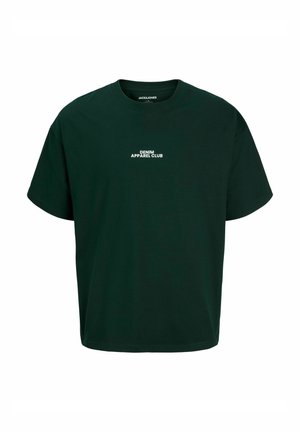 Jeune personne portant un t-shirt vert foncé avec un texte, un jean bleu ample et des chaussures marron, debout devant un fond clair uni.