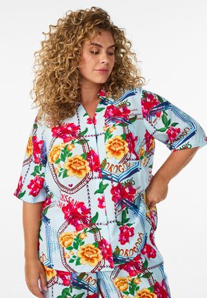 Krøllede kvinde i farverig blomstret og tekst-print kortærmet skjorte og matchende shorts, stående med den ene hånd på hoften mod en hvid baggrund.
