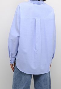 Camicia azzurra a maniche lunghe con colletto, dotata di un dettaglio a pieghe sul retro e polsini con bottoni. Tessuto morbido e liscio con una vestibilità ampia.