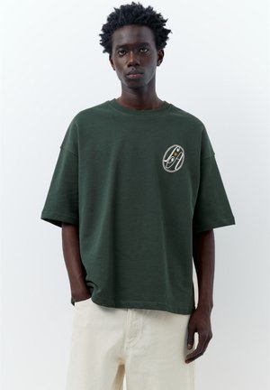 Giovane uomo con pelle scura e capelli ricci che indossa una maglietta verde scuro oversize con un piccolo logo bianco e arancione, in piedi su sfondo bianco.