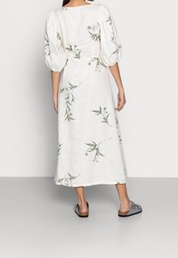 Robe en lin blanc avec motifs floraux verts, manches courtes bouffantes et coupe mi-longue. Portée avec des sandales grises.
