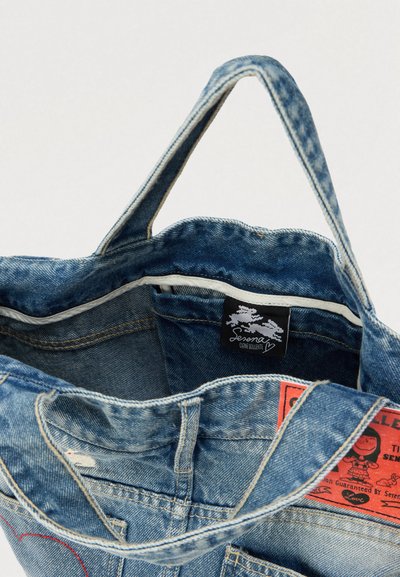 Modrá rifľová taška cez rameno, s bielou podšívkou, spevnenými rukoväťami a vnútornou nálepkou so logom a pokynmi na údržbu.