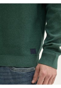 Maglione verde dalla texture a waffle, con polsini e orlo a costine, e una piccola etichetta nera vicino al fondo.