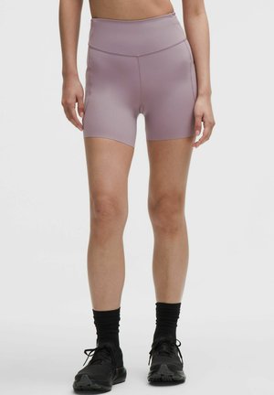 Kobieta w sportowych szortach z wysokim stanem w kolorze mauve, czarnych skarpetach i czarnych butach do biegania, stojąca na tle jasnego, jednolitego tła.