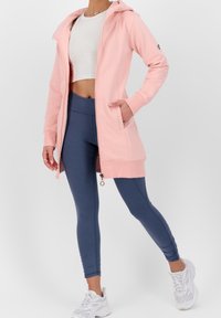 Žena v světle růžové mikině na zip, bílé crop topu, modrých legínách a bílých teniskách, stojí se jednou rukou v kapse.