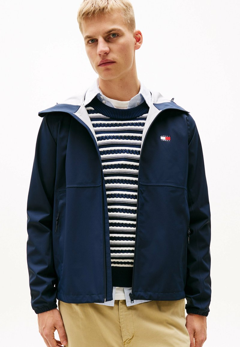 Chaqueta impermeable de color azul marino con capucha gris, bolsillos con cremallera y puños elásticos, llevada sobre un suéter de punto a rayas y una camisa con cuello.