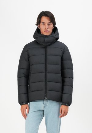 Man wearing black quilted puffer jacket with high collar and light blue jeans, standing against a plain white background.