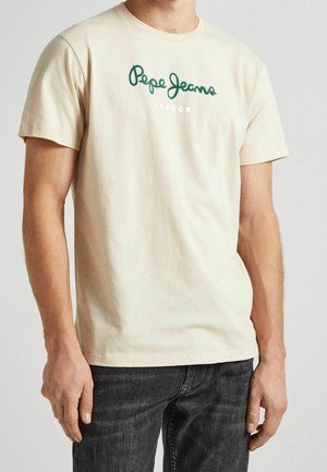 Mężczyzna w kremowym t-shircie z krótkim rękawem z zielonym logo "Pepe Jeans London" i ciemnych dżinsach, stojący z ramionami swobodnie opuszczonymi wzdłuż ciała.