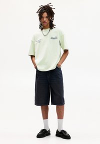 Lys grøn oversized t-shirt med grafiske designs, parret med mørk denim shorts. Sorte sko og hvide sokker fuldender outfittet.