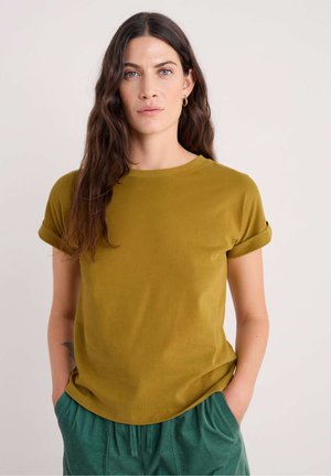 Camicia verde oliva a maniche corte con risvolti, scollatura rotonda e texture liscia. Modello che indossa pantaloni verdi abbinati, mani in tasca.