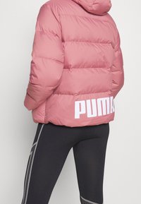 Różowa kurtka puchowa z poziomymi przeszyciami, kapturem i dużym białym logo "PUMA" na plecach. W połączeniu z czarnymi legginsami z bocznymi akcentami.