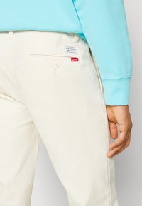 Pantaloni beige chiaro in cotone con una tasca posteriore dotata di chiusura a bottone. Un piccolo etichetta rossa Levi's è visibile sulla tasca.