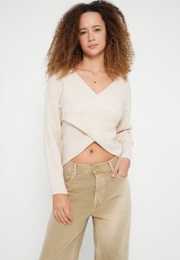 Beige ribgebreide wikkeltrui met een diepe V-hals en lange mouwen, gecombineerd met lichtbeige wijde broekspijpen.